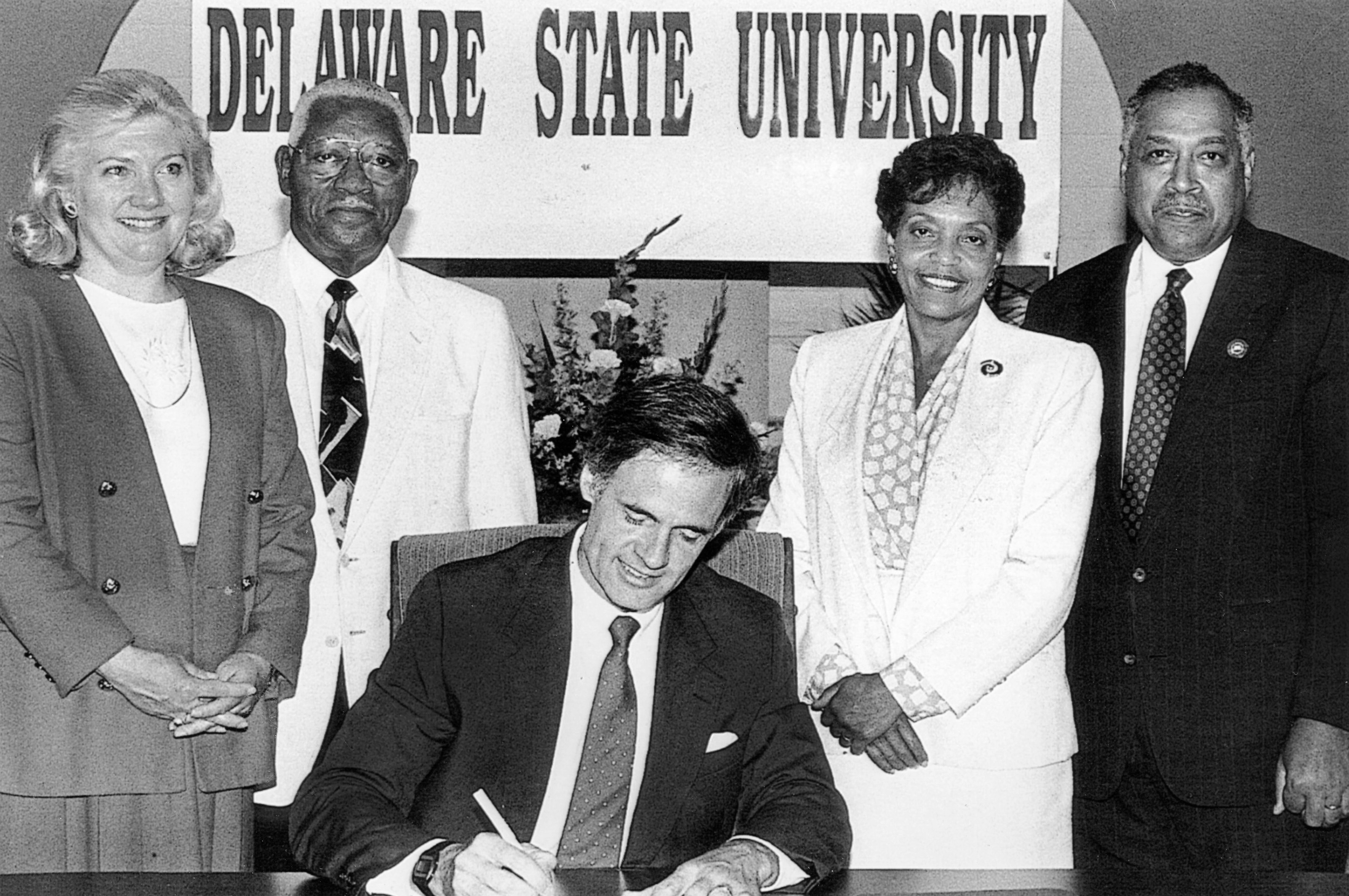 On July 1, 1993, Gov. Tom Carper signs Senate Bill 138, which changed Delaware State College to Delaware State University. Looking on, from left, are Representative Nancy Wagner, Senator Herman Holloway Sr., Vermell DeLauder and President William B. DeLauder.