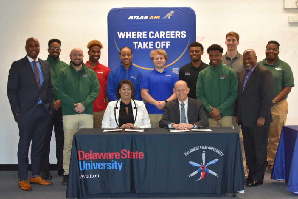 College of Business Dean Michael Casson, Aviation Program Director Lt. Col. Michael Hales (both standing in suit jackets) and a group of DSU Aviation students flanks the seated Jackie Griffith, DSU Chief of Staff and Capt. Jeff Carlson, Atlas Air Sr. VP of Flight Operation, after the two signed a new agreement that establishes the air provider's University Pathway to Success Program at DSU, which will enable Aviation graduates to pursue a direct career path of flying Boeing 737 as a First Officer early in their career.