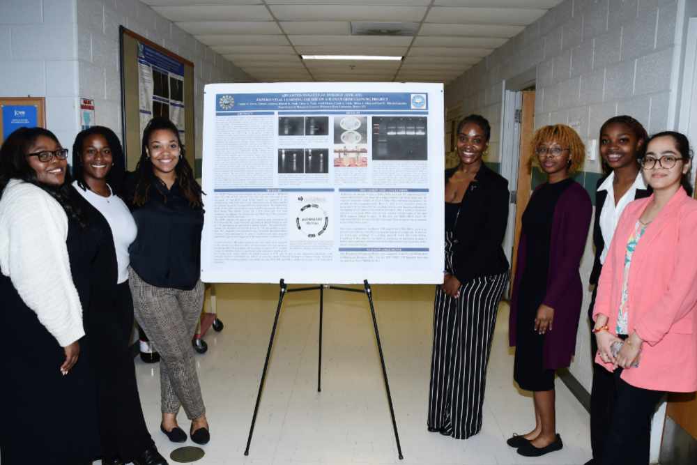 (L-r) Asiah Green, Tionah Johnson, Taylor Davis, Milan King, Calah Vicks, Chloe Tuck, and Khushi Shah stand with the research poster they jointly produced in their Experiential Learning Course on a Human Gene Cloning Project.